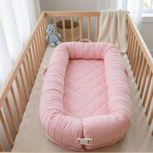 Pink baby nest in a crib with a teddy bear and blanket in the background, baby nest, baby nest bed, newborn lounger, infant lounger, cotton baby nest, portable baby lounger, baby nest with removable cover, baby lounger washable, newborn gift, baby shower gift, nursery essential